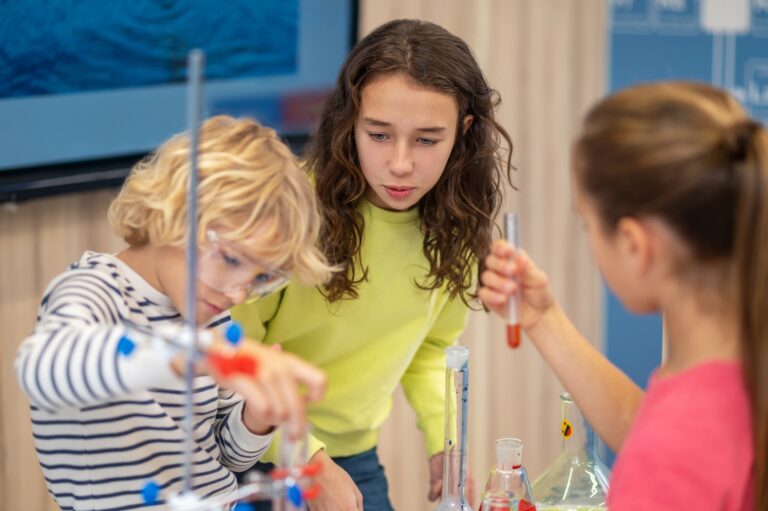Practical chemistry. Engaged surprised girl watching with interest classmates with test tubes standing near table in chemistry class