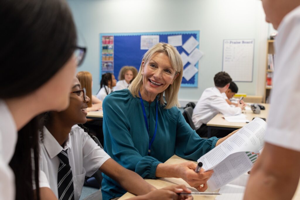 Close-up shot of a small group of students and their teacher sitting in a classroom in a school in Newcastle upon Tyne, North East England. The main focus is on the smiling teacher who is showing the students a worksheet. The students are wearing school uniforms.Videos similar to this scenario available.
